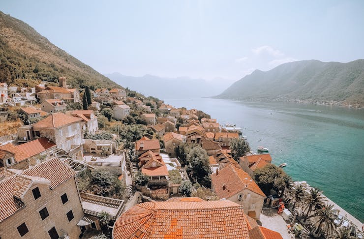 an aerial shot of a mountainous cliffside dotted with medieval-style houses overlooking a beautiful turquoise bay