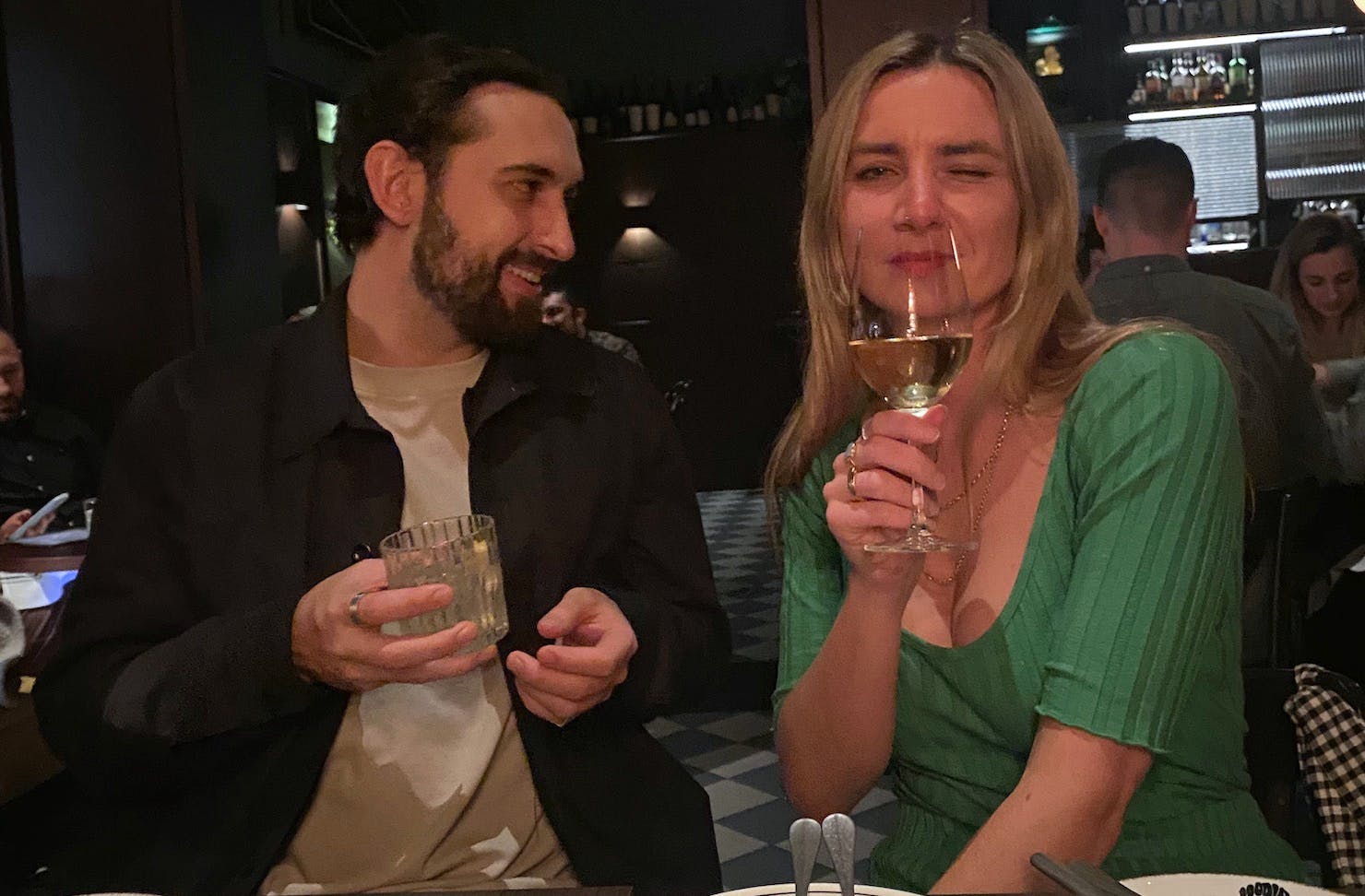 A man and woman sitting in a moody restaurant hold their drinks. The woman winks at the camera.
