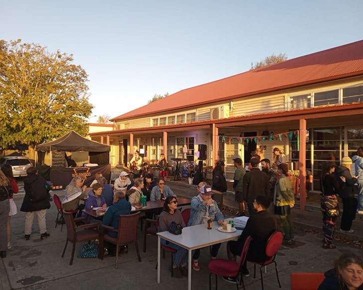 People sit around tables at the Vegan Night MÄkete as the sun sets, lighting up buildings in gold.