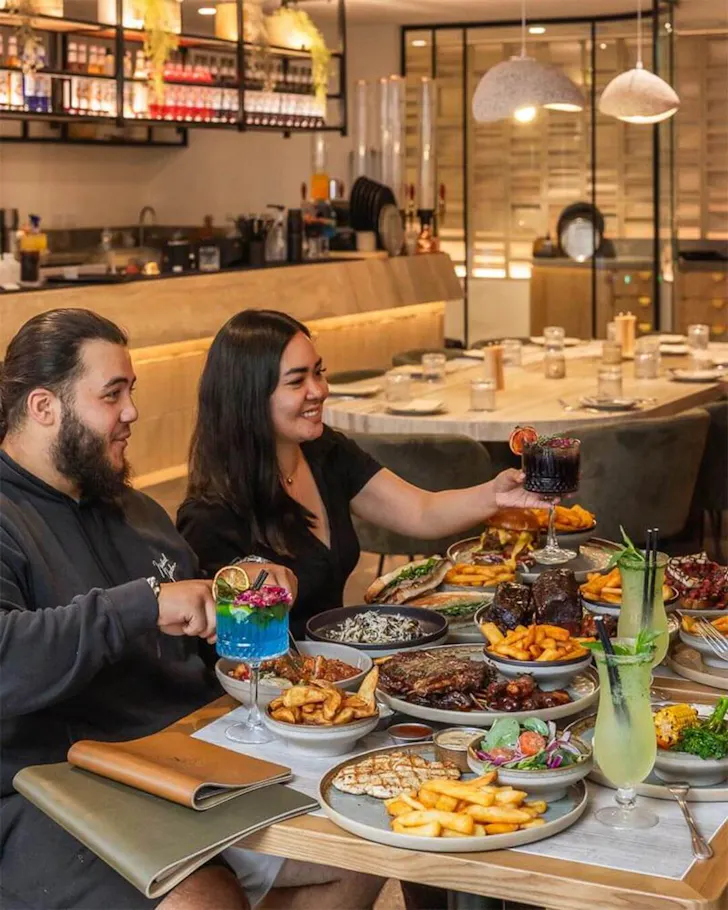 A mid-shot of a couple having dinner at a table, with the rest of the restaurant and bar in the background. The couple are shown in the left side of the image at the forefront, eating from a table of many dishes of meats, salads, chips and a few colourful cocktail drinks.