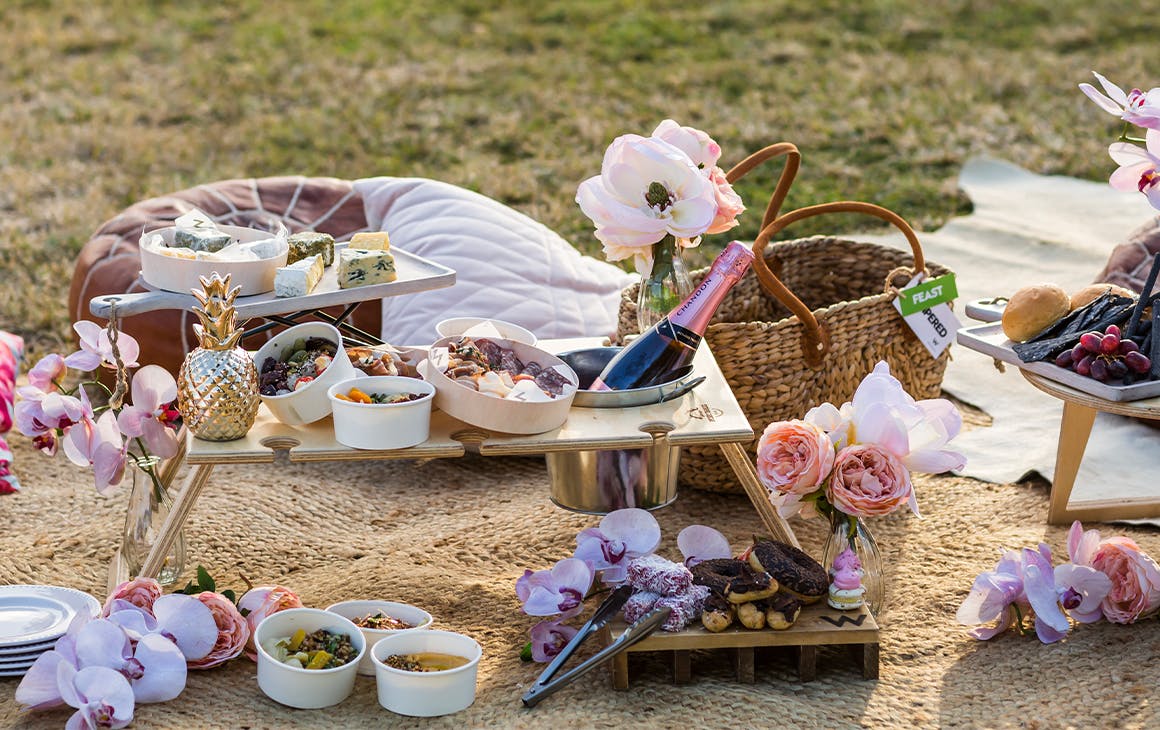 A display of food and beverages from a picnic hamper on a blanket over the grass