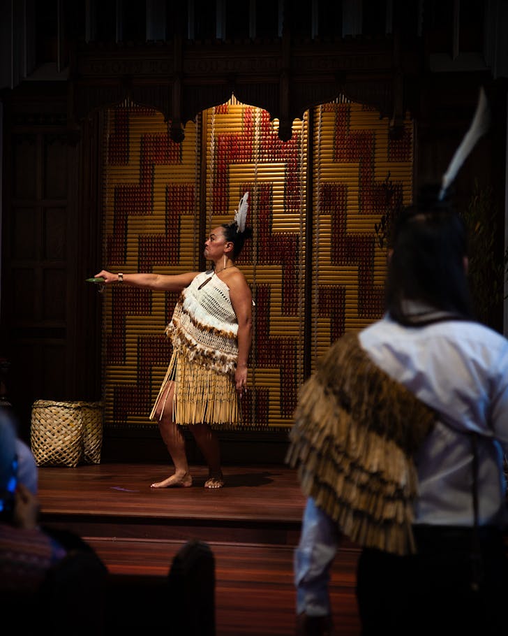 A woman in traditional MÄori attire stands, holding out a carved ponamu.