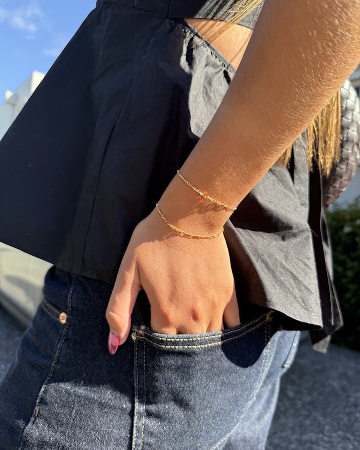 A close-up of someone standing outside on a sunny day in a cute black top and dark blue jeans, with their hand in their back pocket, shows some gold jewellery from the Welder's Daughter on their wrist.