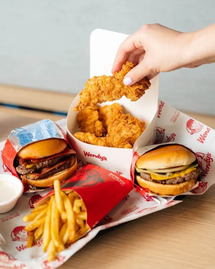 Wendy's meal featuring two cheeseburgers, fries and crispy chicken tenders, with a hand reaching into the box to pick up a chicken tender.