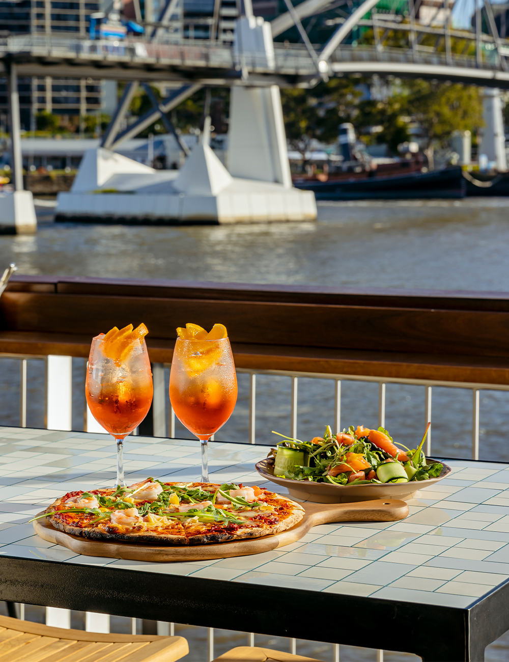 two cocktails and a pizza on a table overlooking the river