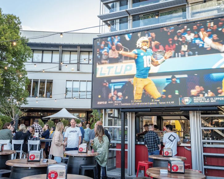 A wide shot of an outdoor pub seating area with a few people mingling while a giant screen plays a live sport game.