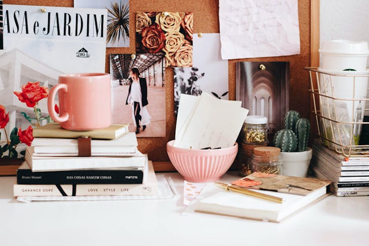 A desk with a pink mug, scattered books, and a pin board. 