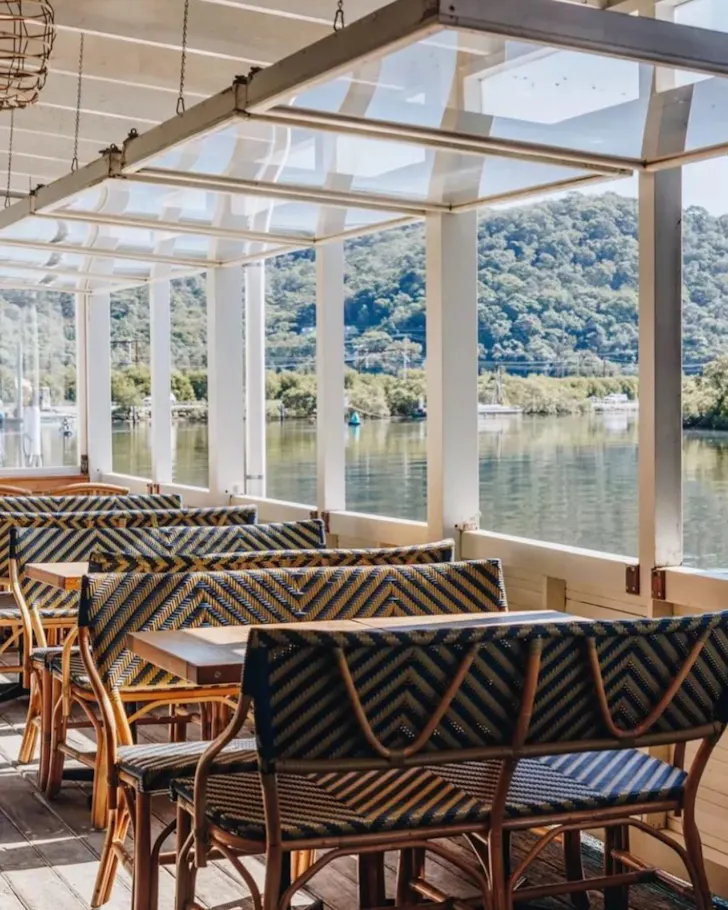 Covered waterfront dining terrace with patterned wicker seating overlooking a calm river framed by lush, tree-covered hills on the Central Coast.