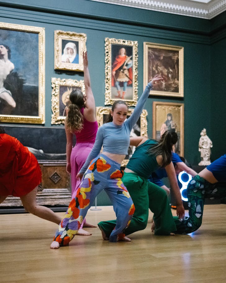 Young dancers strike a pose/execute a routine in front of a bluish gallery wall covered in grand paintings.