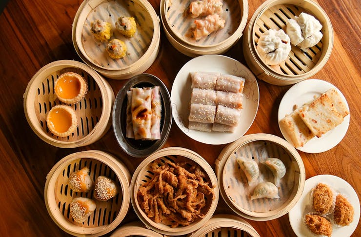 A selection yum cha dishes like pork buns, dumplings and egg rolls in bamboo baskets spread across a table.