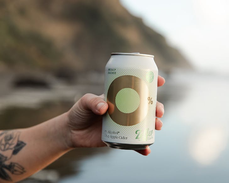Someone holds a can of cider out with a wild beach in the background.