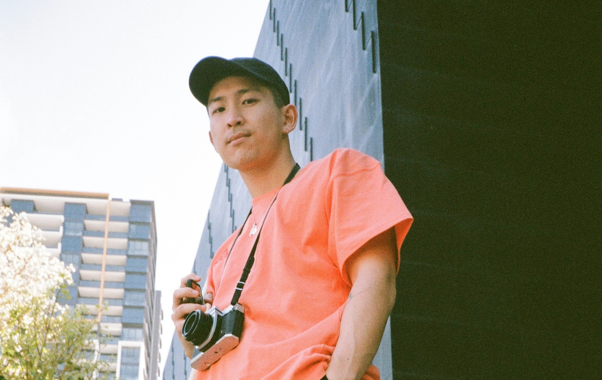 A person in an orange t-shirt looking down at the camera, and also holding a camera in their hand.