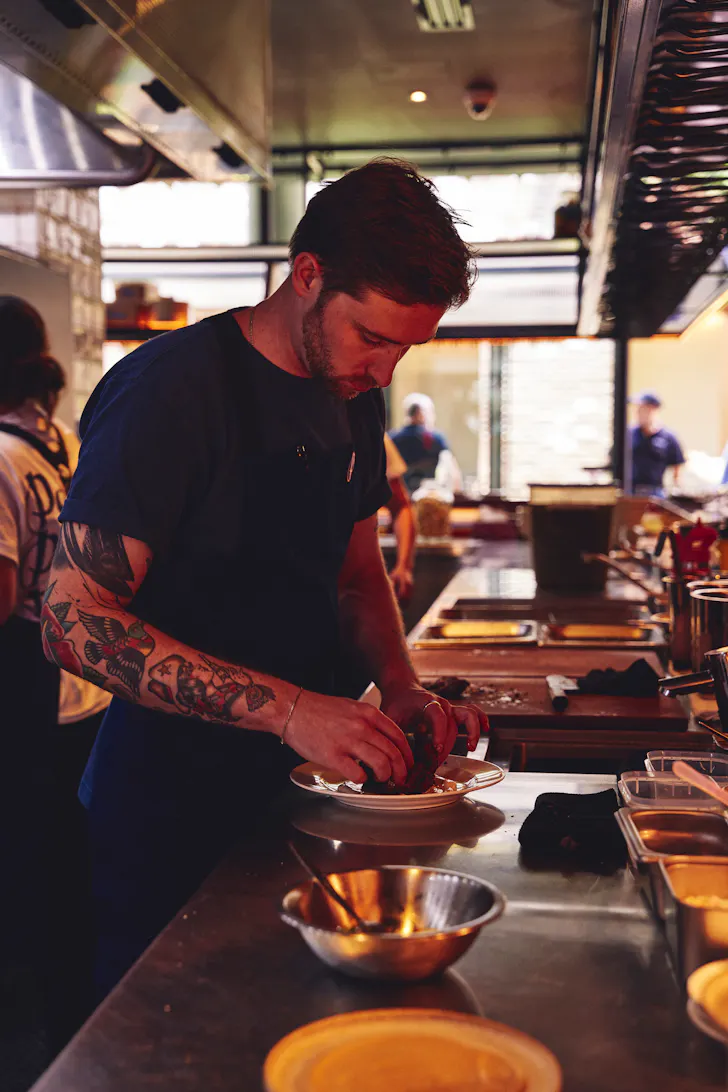 A bustling open kitchen with chefs moving through service unfolds against exposed brick walls, capturing the energy and warmth of a busy restaurant.