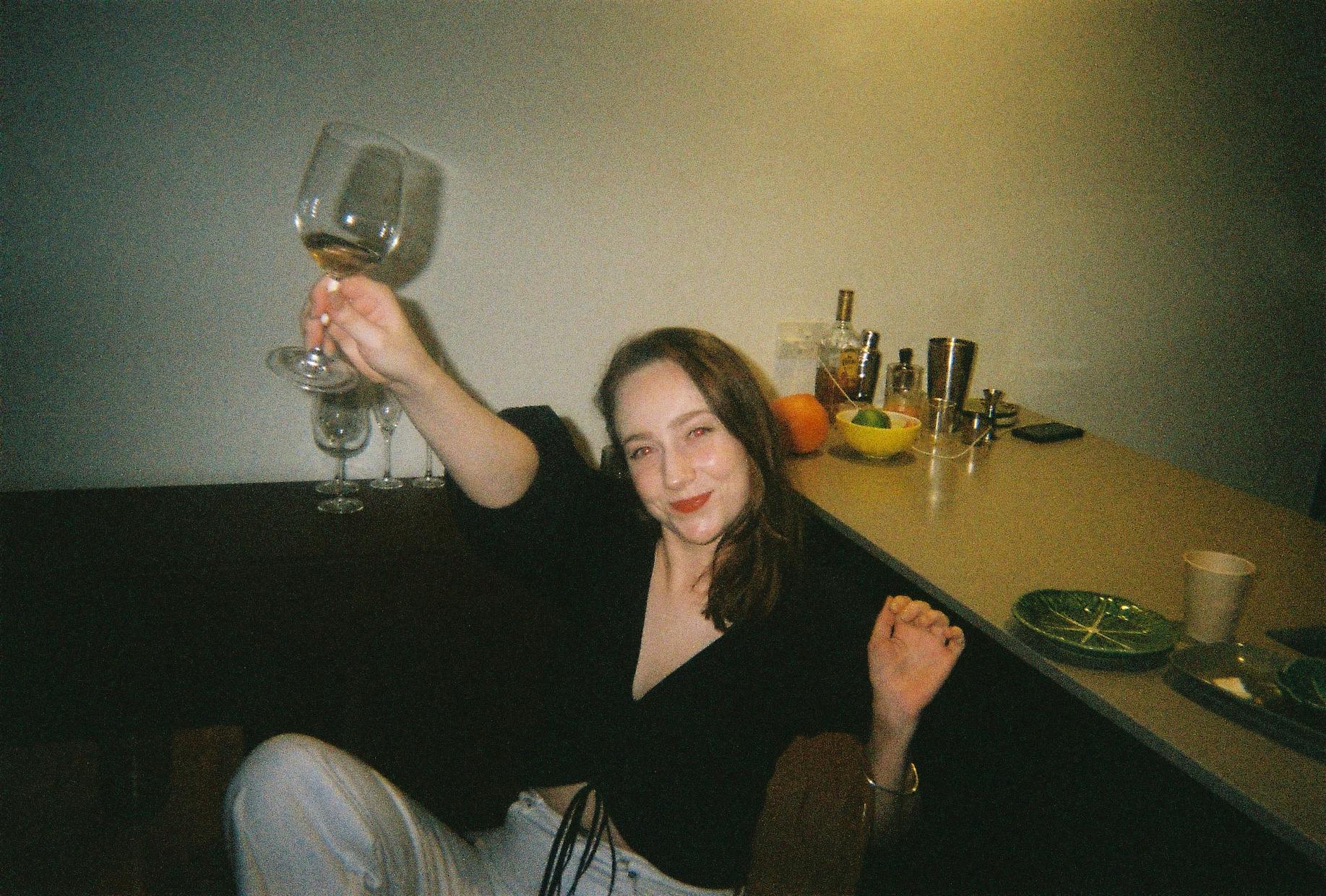 A person in the kitchen joyously holding up a wine glass with a small amount of wine in it. 