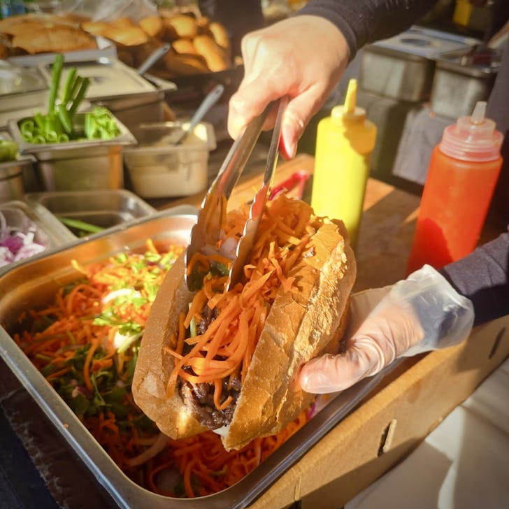 a hand holding out a banh mi and putting veggies on it