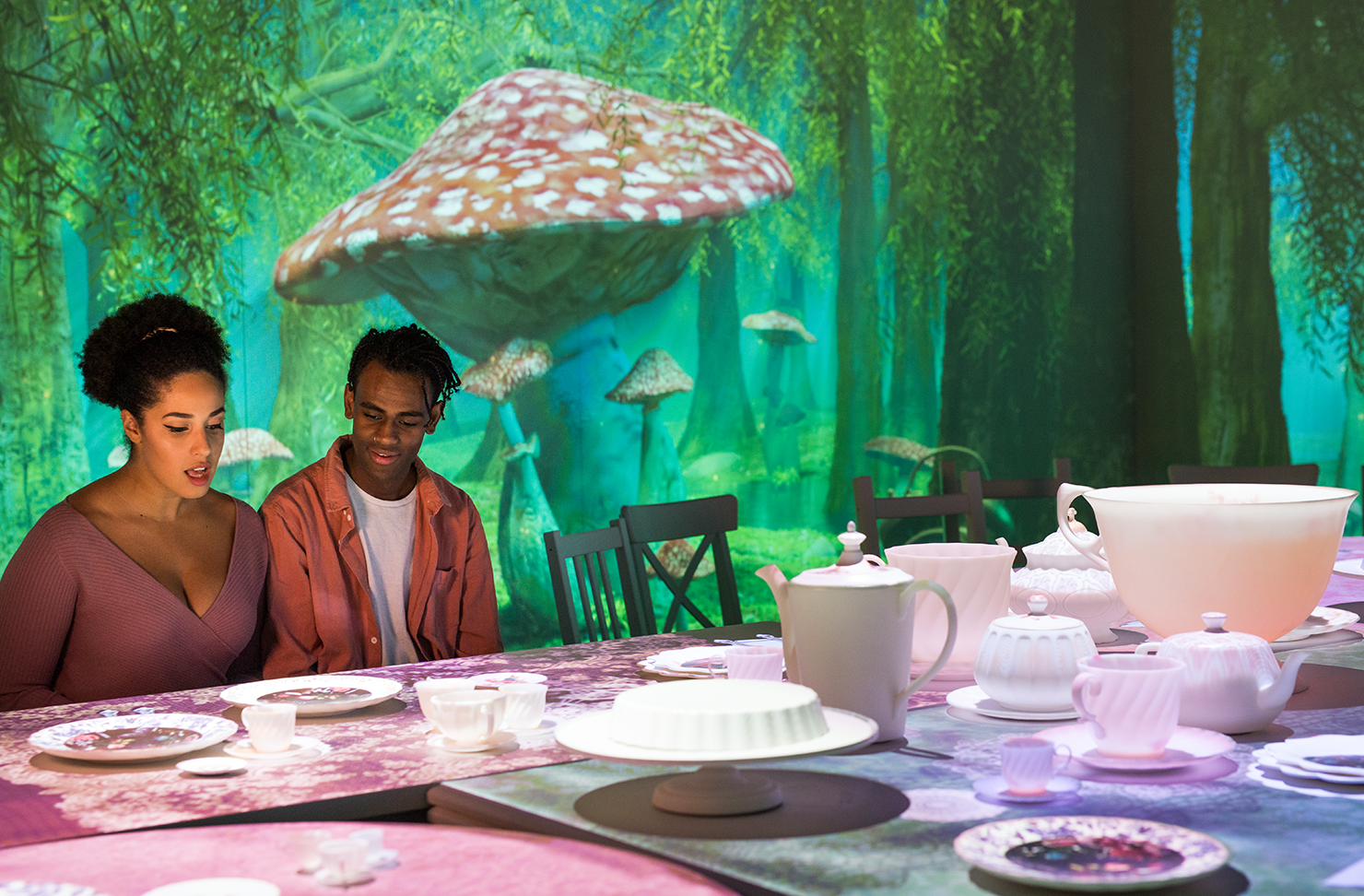 Two people sitting at the Mad Hatter's Tea Party with a large mushroom behind them.