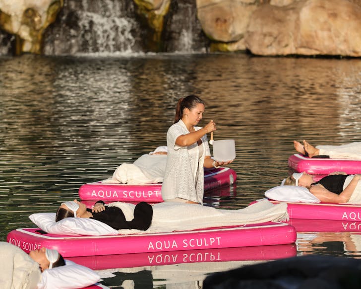 People floating atop pink blow up mattresses with white blankets, pillows and eye masks on a saltwater pool while a lady walks between them with a singing bowl