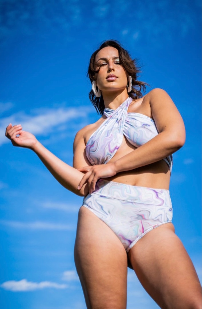 A woman stands against the blue sky wearing a hippy print bikini with large shell hoop earrings. 
