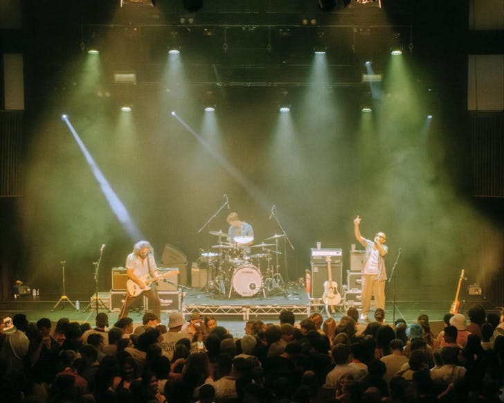 Three males - a drummer, guitarist and singer - perform on stage in front of a large crowd.