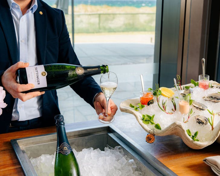 Man in suit is pouring champagne into a flute next to a large, ice-filled shell scattered with oysters and jars of oyster toppings.