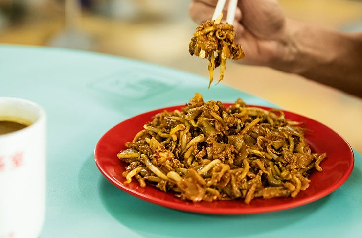 A plate of Char Kway Teow on a blue table with a person's hand using chopsticks.