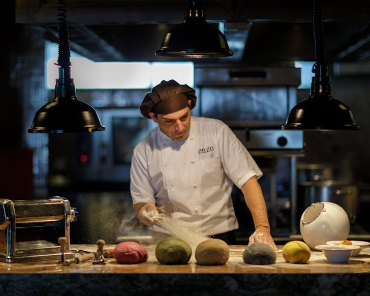 Chef Enzo wearing brown chefs hat and white chefs shirt in a dark kitchen, throwing flour across dough