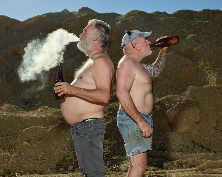 Two older caucasian men stand back to back drinking beers and smoking cigarettes with no shirts on in front of distant mountains.