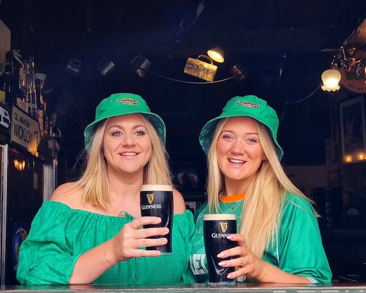 Two caucasian women with blonde hair sit in a pub wearing green St Patrick's Day hats and cheersing two pints of Guiness