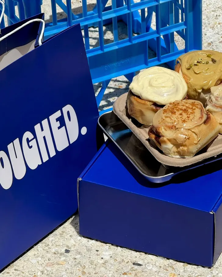 A tray of cinnamon scrolls sit on a blue crate next to a blue Doughed takeaway bag