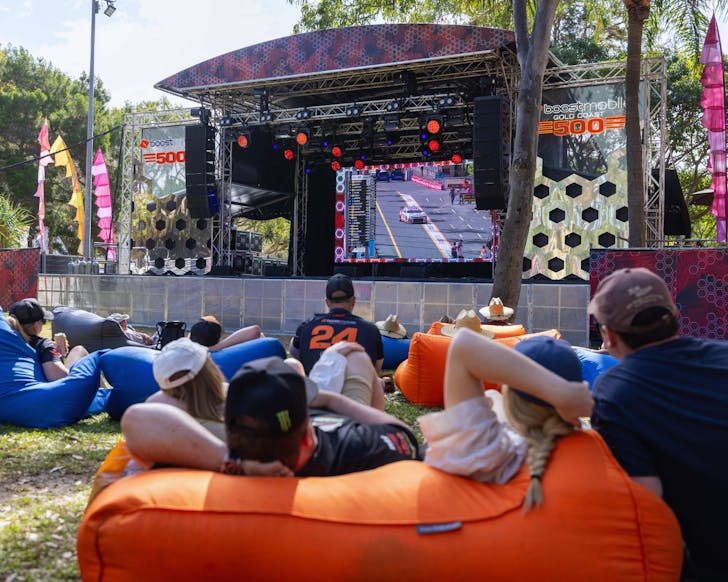 Groups of people sit on orange bean bags on the grass, watching the GC500 race on a large screen.