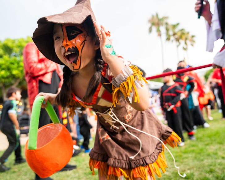 A small female child wearing a brown and orange witches costume and spooky pumpkin face paint runs through a park and groups of other dressed up children. She is carrying a pumpkin-themed treat basket.