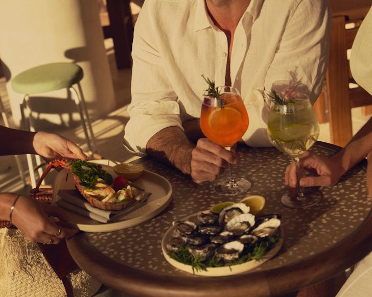 Two people sitting at a table holding an orange spritz and a slightly green spritz, being served plates of oysters and a lobster dish