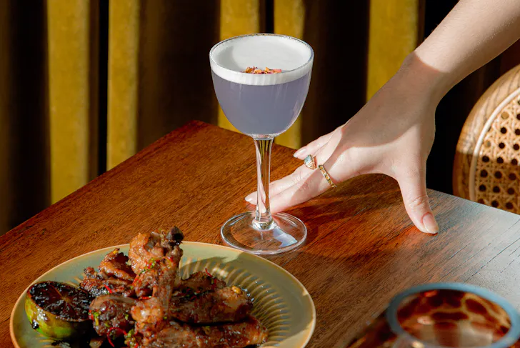 A purple cocktail and food sit on a timber table, with a hand resting next to them and olive green curtain in the background at Maggie May, Gasworks