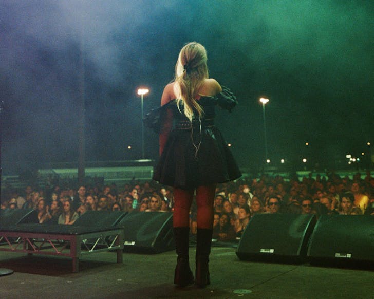 Blonde haired female stands centre stage, facing crowd. Image is taken from behind her, looking out at crowd. She wears a short, black bubble dress and chunky black ankle boots.