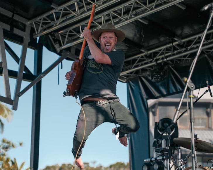 Caucasian man is leaping in the air on stage playing the guitar.