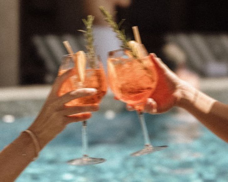 Two orange Aperol spritz cocktails clinking together in a cheersing motion in front of a pool.