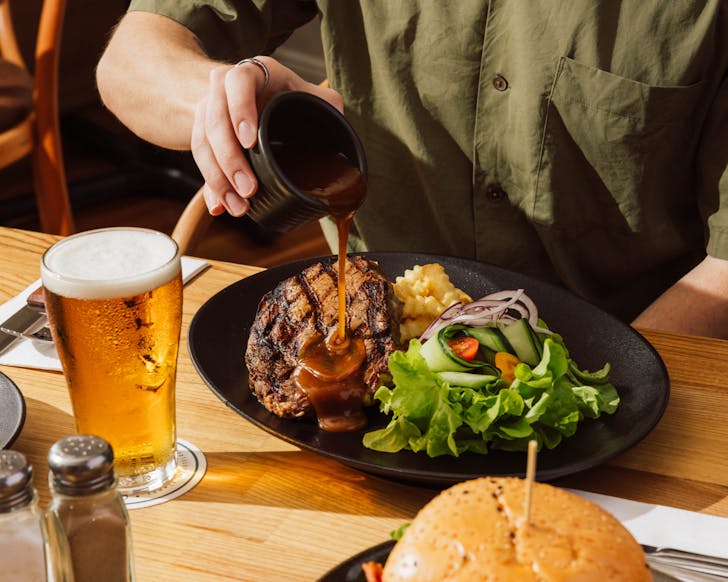 Hand pours gravy looking sauce over the top of a steak, which is sitting on a plate with a salad and some crinkle cut potato chips. There is a beer sitting next to the plate on the table.