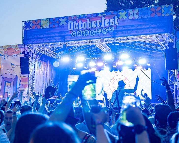 A crowd dressed in Oktoberfest wear dances in front of a stage, which is topped with a large banner reading Oktoberfest In The Gardens.