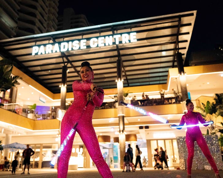 Female dancers in sparkling hot pink outfits twirl LED lights in front of Paradise Centre sign at night