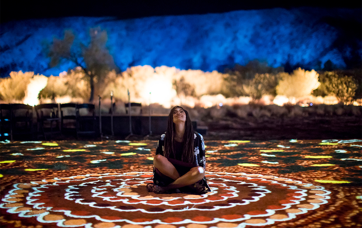 a young girl sits on the ground - which is lit up in various colours - at Alice Springs Desert Park