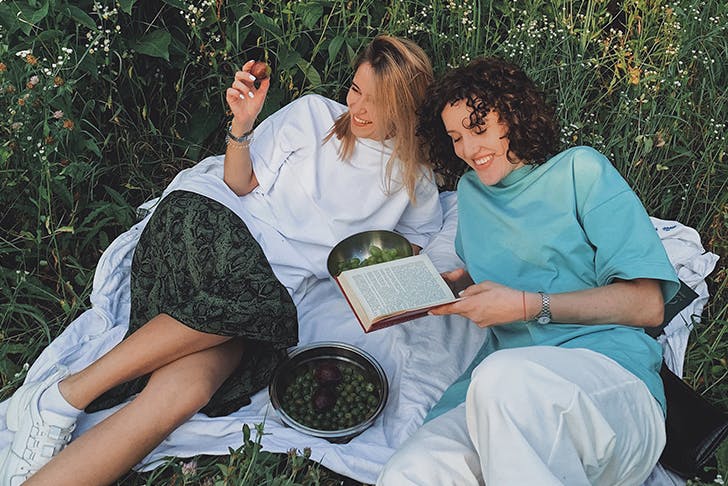 Two women laying on a picnic blanket in the grass laughing, holding a book and eating olives.