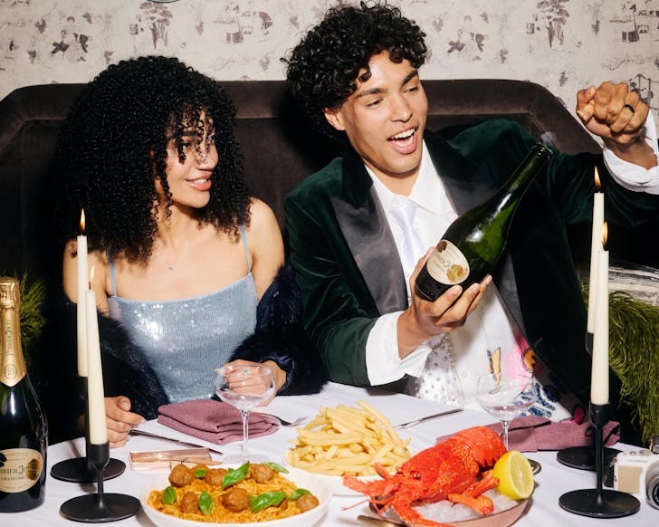 A male and a female sit behind a table covered with plates of chips, lobster, spaghetti and meatballs, and long candle sticks, popping a bottle of champagne.