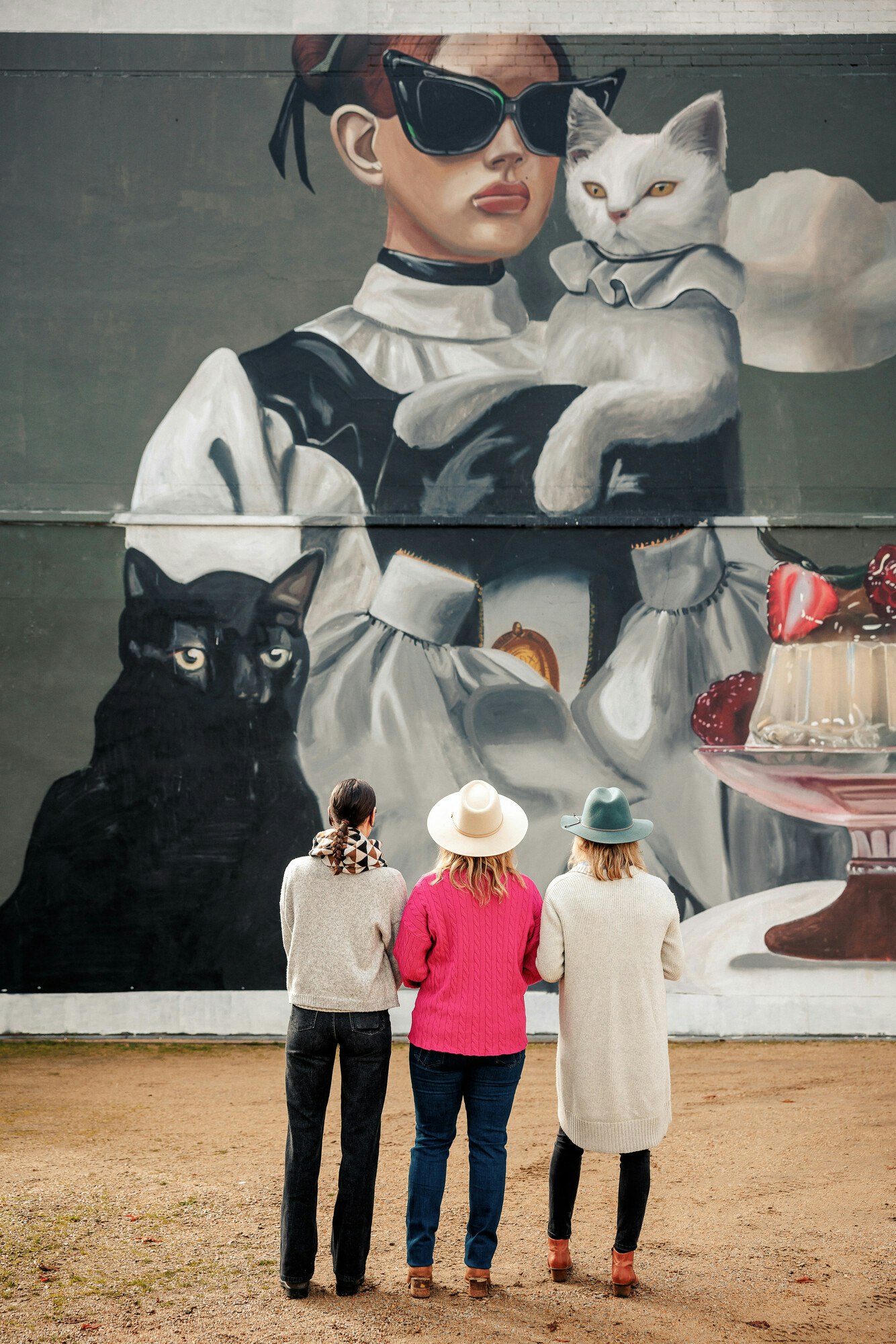 Three people admiring a mural of a woman with cats and desserts