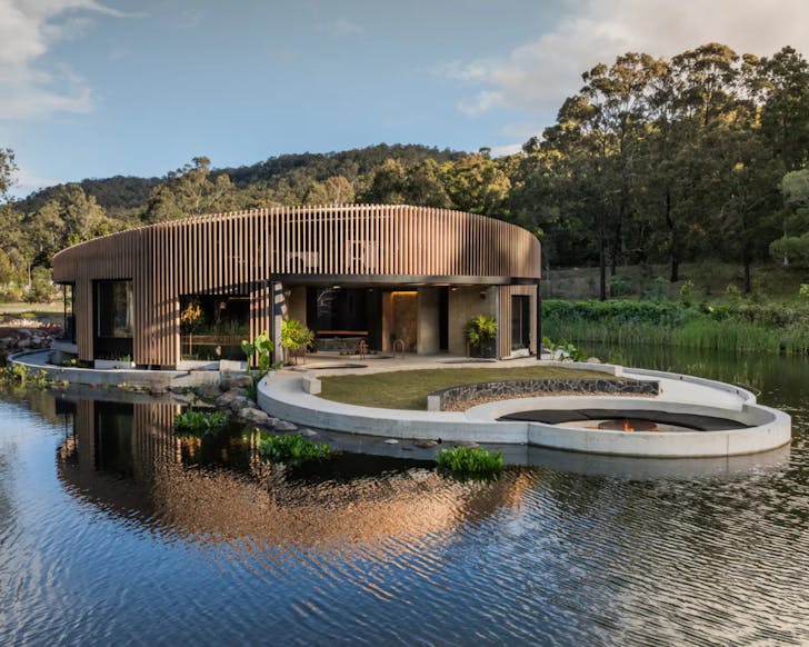 Circular bathhouse with warm, wooden exterior and large rectangular windows perched upon a lake under a blue sky.
