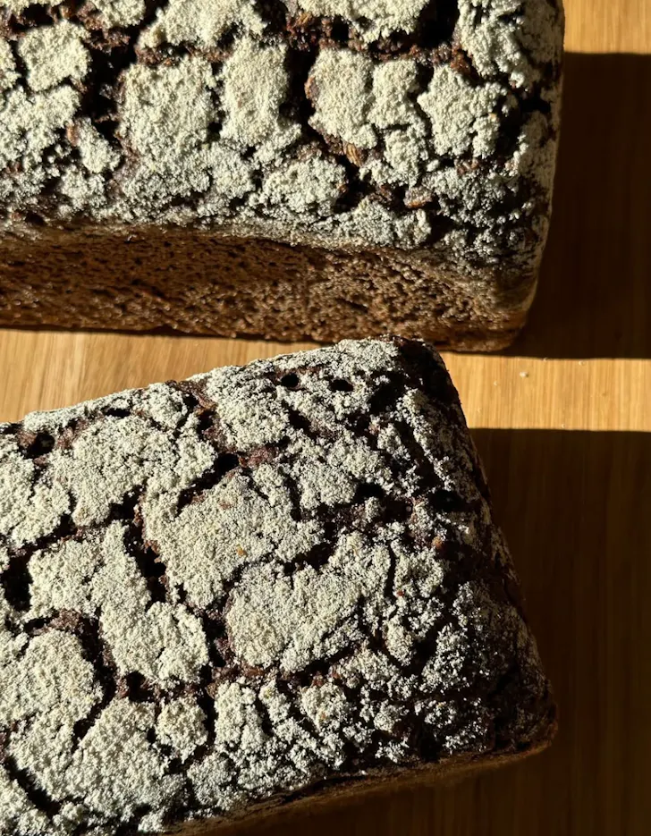 Two dark rye loaves with a heavily floured, crackled crust sit in warm natural light on a wooden surface, highlighting their rustic texture.