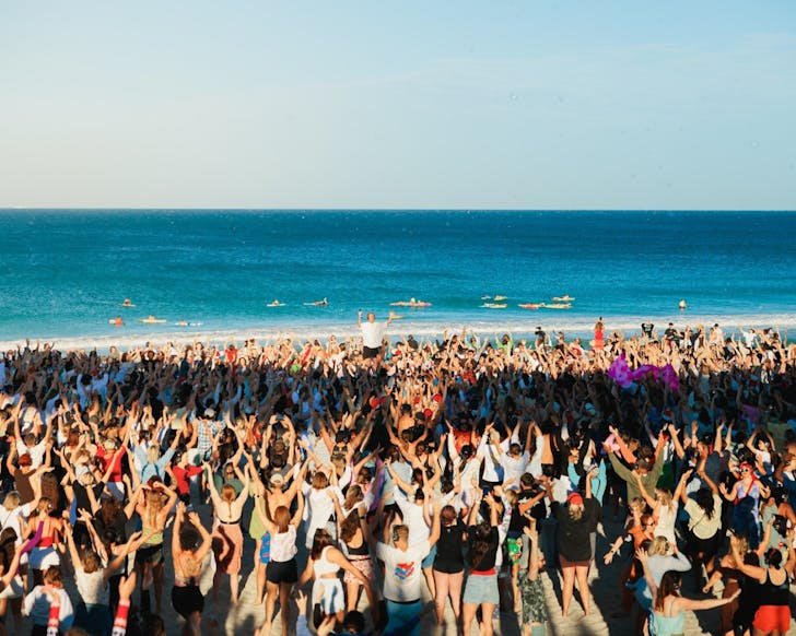 A large group of people dancing with headphones on at the beach at a silent disco. There are surfers in the ocean behind them.