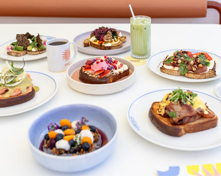 Scattered colourful plates of breakfast dishes such as French toast, eggs Benedict, blueberry oats, mushroom and eggs on toast, smoked salmon topped with a green sauce on toast, and scattered coffees and an iced matcha, on a white table. 