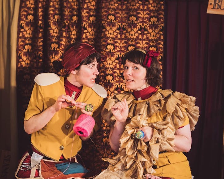 Two seamstresses wearing warm coloured eccentric outfits that they've seemingly sewn and stitched themselves, surrounded by sewing tools such as scissors and string 