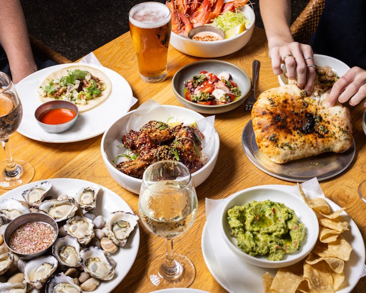 Table covered in various dishes: plate of oysters, plate of corn chips and guacamole, king prawns, sticky chicken wings, tacos and flatbread being torn by a pair of hands. There are scattered glasses of white wine and beer between the dishes.