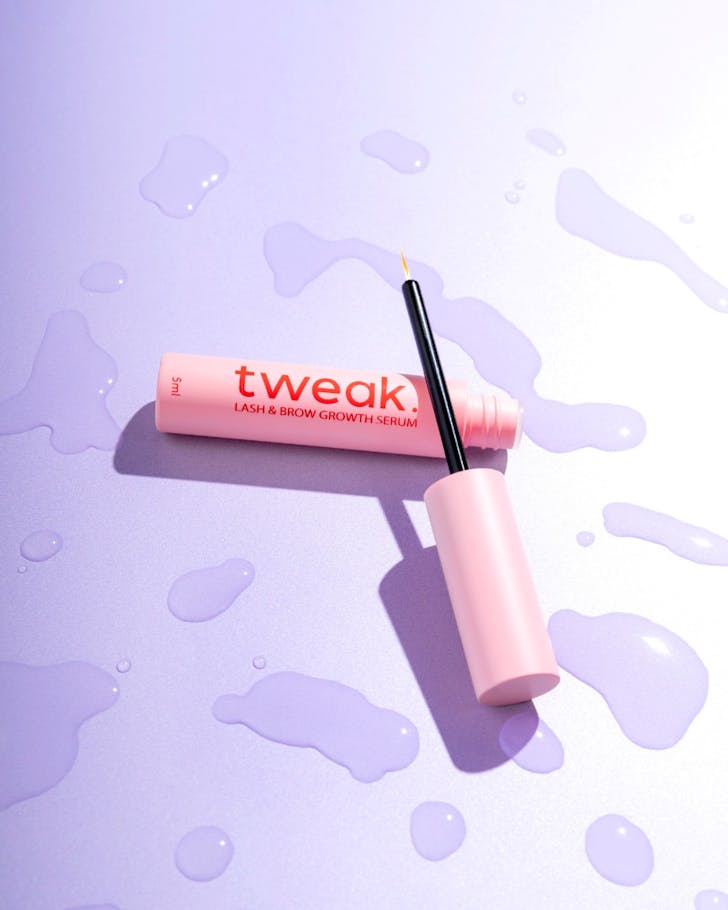 A pink Tweak bottle with the wand lying next to it sits on a purple background with water spots surrounding them
